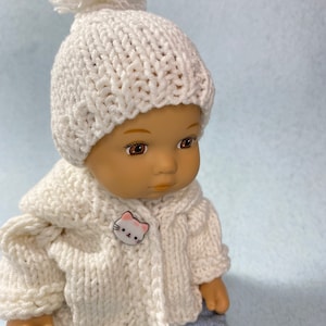 May include: A white knitted doll hat with a white pom pom on top. The doll is wearing a white knitted cardigan with a white cat button. The doll is wearing blue jeans.