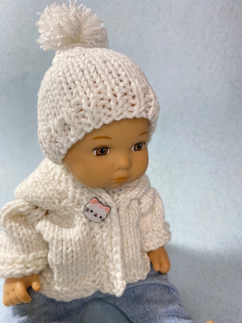 May include: A white knitted doll hat with a white pom pom on top. The doll is wearing a white knitted cardigan with a white cat button. The doll is wearing blue jeans.