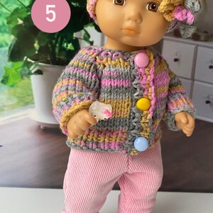 Puede incluir: Una mu&ntilde;eca con un c&aacute;rdigan de punto a rayas grises, rosas y amarillas con botones de colores, pantalones rosas y zapatillas blancas. La mu&ntilde;eca lleva un gorro de punto gris, rosa y amarillo con una flor.