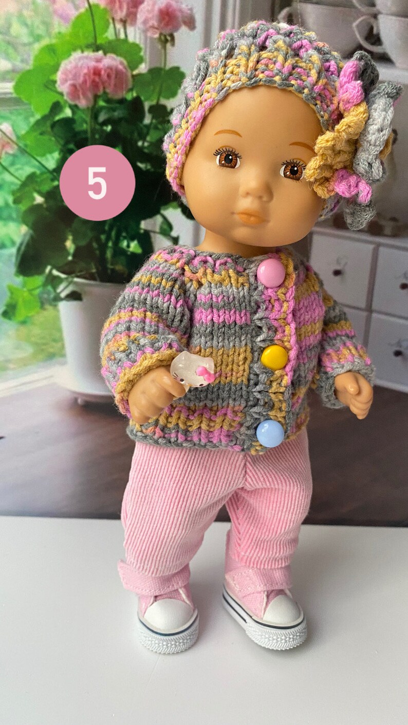 Puede incluir: Una mu&ntilde;eca con un c&aacute;rdigan de punto a rayas grises, rosas y amarillas con botones de colores, pantalones rosas y zapatillas blancas. La mu&ntilde;eca lleva un gorro de punto gris, rosa y amarillo con una flor.