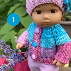 Puede incluir: Una mu&ntilde;eca de punto que lleva un su&eacute;ter a rayas rosa y azul, un gorro rosa con una flor y pantalones rosas. La mu&ntilde;eca lleva zapatillas blancas y est&aacute; de pie sobre una superficie de madera.