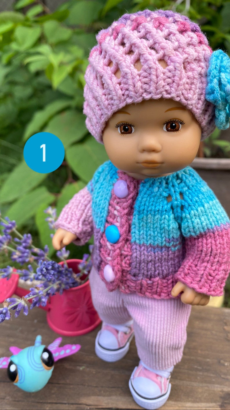 Puede incluir: Una mu&ntilde;eca de punto que lleva un su&eacute;ter a rayas rosa y azul, un gorro rosa con una flor y pantalones rosas. La mu&ntilde;eca lleva zapatillas blancas y est&aacute; de pie sobre una superficie de madera.