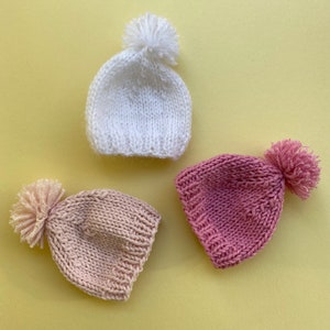 May include: Three knitted hats with pom-poms in white, pink, and light pink.