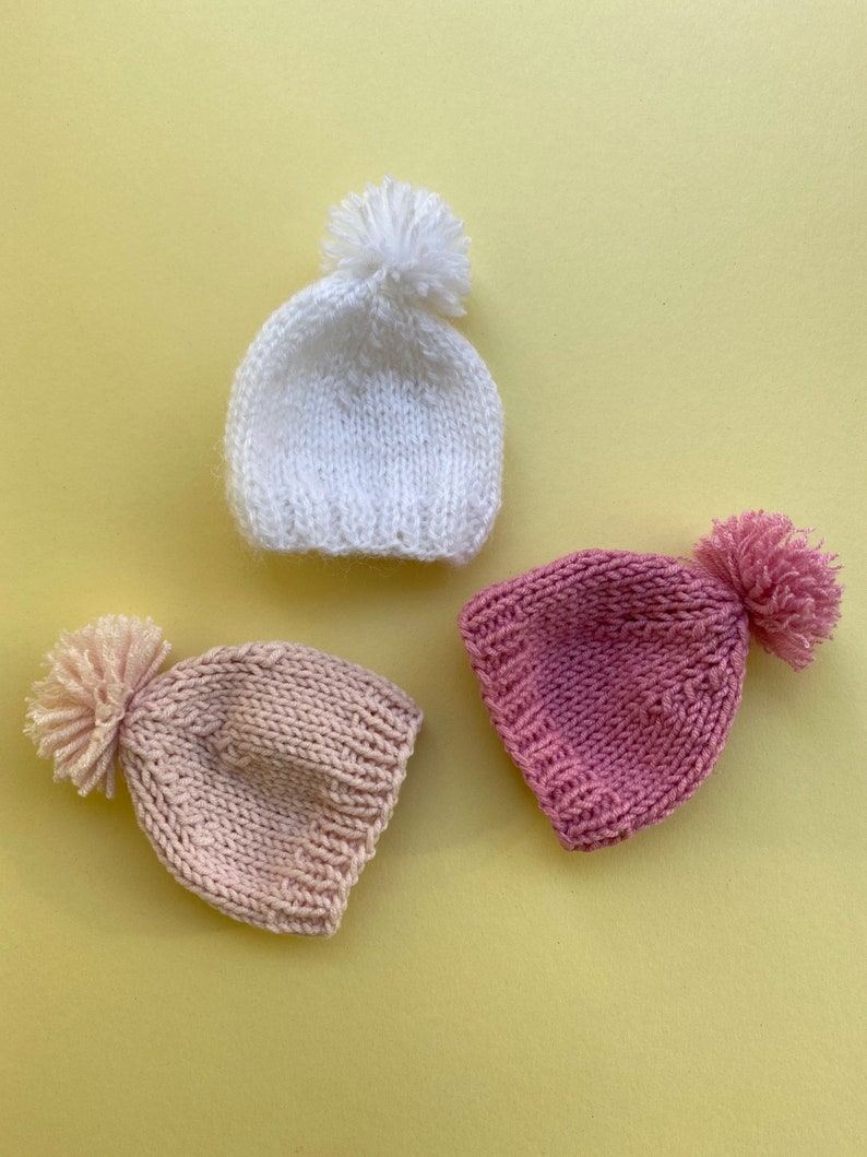 May include: Three knitted hats with pom-poms in white, pink, and light pink.