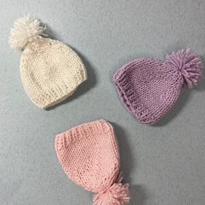 May include: Three knitted hats with pom-poms. One hat is white, one is pink, and one is purple.