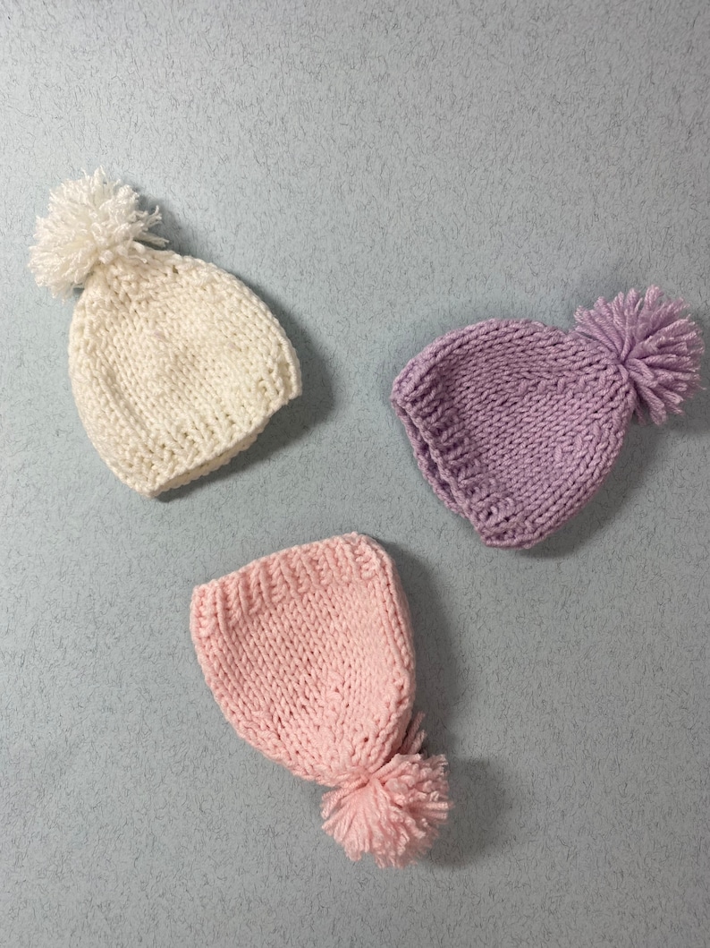 May include: Three knitted hats with pom-poms. One hat is white, one is pink, and one is purple.