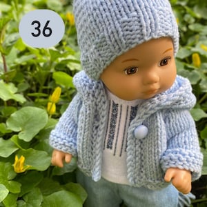 May include: A blue knitted doll outfit with a pom-pom hat, cardigan, and pants. The doll is wearing a white shirt with blue embroidery. The doll is standing in a bed of green leaves and yellow flowers.
