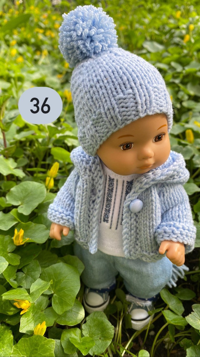 May include: A blue knitted doll outfit with a pom-pom hat, cardigan, and pants. The doll is wearing a white shirt with blue embroidery. The doll is standing in a bed of green leaves and yellow flowers.