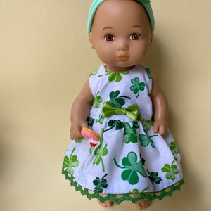 May include: A doll wearing a white dress with green shamrock print and a green headband with a shamrock hat. The doll is holding a pink and white donut.