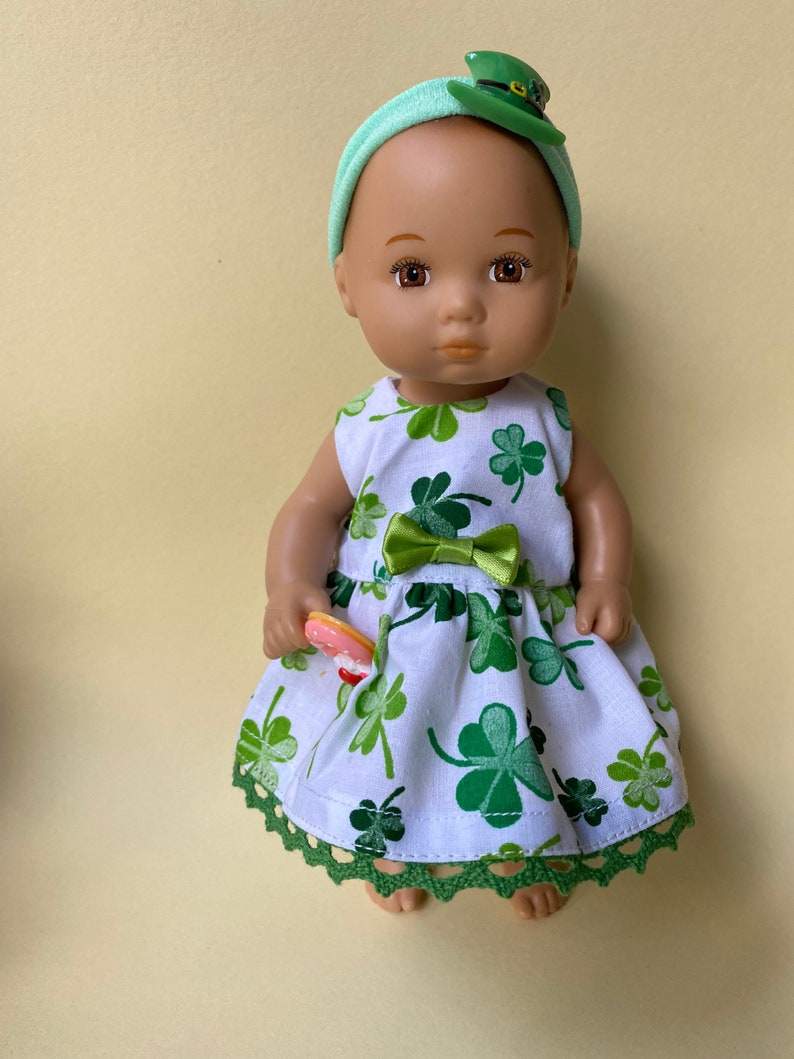 May include: A doll wearing a white dress with green shamrock print and a green headband with a shamrock hat. The doll is holding a pink and white donut.