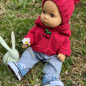 May include: A red knitted doll with a cherry design on its sweater, wearing blue jeans and blue sneakers, sitting on green grass.