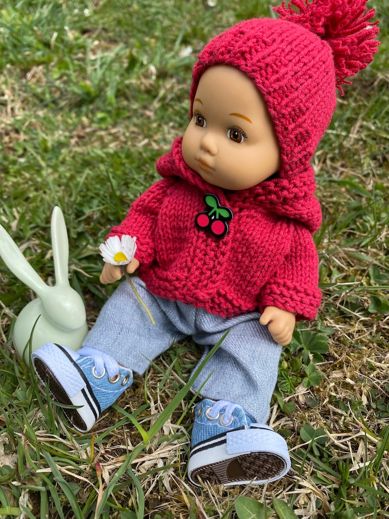 May include: A red knitted doll with a cherry design on its sweater, wearing blue jeans and blue sneakers, sitting on green grass.