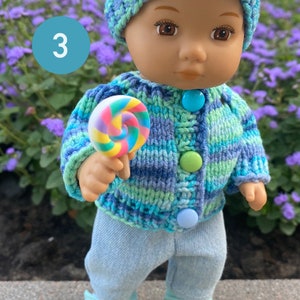 Puede incluir: Una mu&ntilde;eca con un su&eacute;ter de punto a rayas azul y verde con botones azules, pantalones vaqueros azules y un gorro de punto azul y verde. La mu&ntilde;eca sostiene una piruleta colorida.