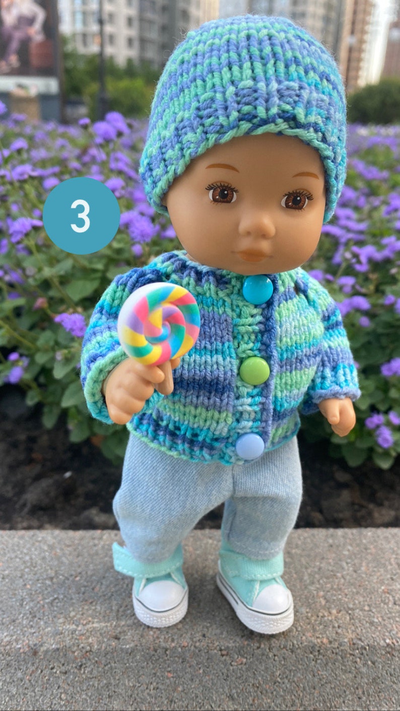 Puede incluir: Una mu&ntilde;eca con un su&eacute;ter de punto a rayas azul y verde con botones azules, pantalones vaqueros azules y un gorro de punto azul y verde. La mu&ntilde;eca sostiene una piruleta colorida.
