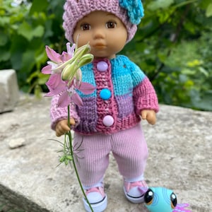 Puede incluir: Una mu&ntilde;eca con un c&aacute;rdigan de punto rosa y azul, pantalones rosas y un gorro de punto rosa con una flor azul. La mu&ntilde;eca sostiene una flor rosa y est&aacute; de pie sobre una superficie de piedra con un juguete azul y morado.