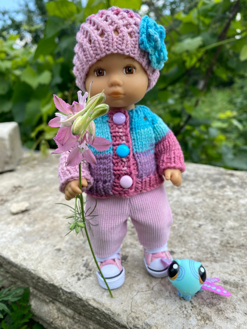 Puede incluir: Una mu&ntilde;eca con un c&aacute;rdigan de punto rosa y azul, pantalones rosas y un gorro de punto rosa con una flor azul. La mu&ntilde;eca sostiene una flor rosa y est&aacute; de pie sobre una superficie de piedra con un juguete azul y morado.