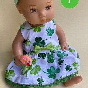 May include: A doll wearing a green shamrock print dress with a green bow, a green headband with a green hat, and holding a pink and white donut. The doll has brown eyes and brown hair. The background is a light yellow.