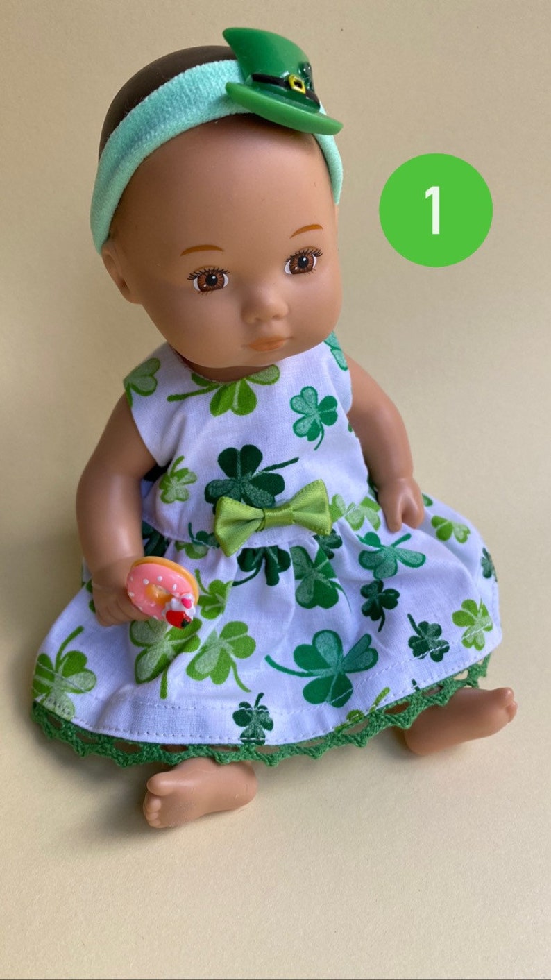 May include: A doll wearing a green shamrock print dress with a green bow, a green headband with a green hat, and holding a pink and white donut. The doll has brown eyes and brown hair. The background is a light yellow.
