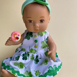 May include: A doll wearing a white dress with green shamrock print and a green headband with a green shamrock hat. The doll is holding a pink and white donut.