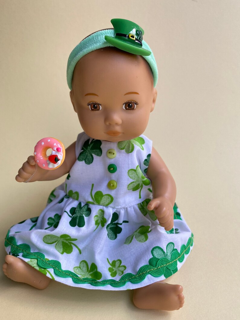 May include: A doll wearing a white dress with green shamrock print and a green headband with a green shamrock hat. The doll is holding a pink and white donut.