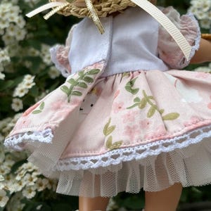 May include: A doll wearing a straw hat with a pink flower and ribbon, a white top, and a pink floral print dress with white lace trim. The doll is also wearing white shoes.