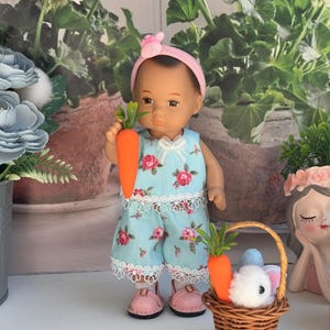 May include: A doll wearing a blue floral dress and a pink headband with bunny ears holds a carrot. A wicker basket with a white bunny and a carrot sits in front of the doll.