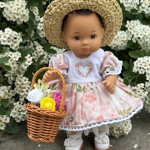 May include: A doll wearing a straw hat, a pink floral dress, and white shoes with bows. The doll is holding a small woven basket filled with colorful flowers. The doll is standing in front of a backdrop of white flowers.