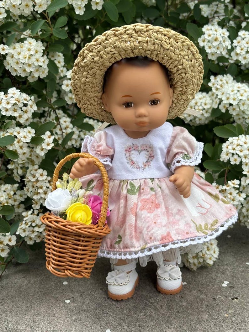 May include: A doll wearing a straw hat, a pink floral dress, and white shoes with bows. The doll is holding a small woven basket filled with colorful flowers. The doll is standing in front of a backdrop of white flowers.