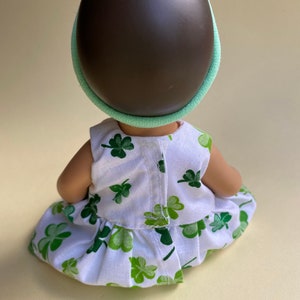 May include: A small doll wearing a white dress with green shamrock print and a green headband with a shamrock on top.