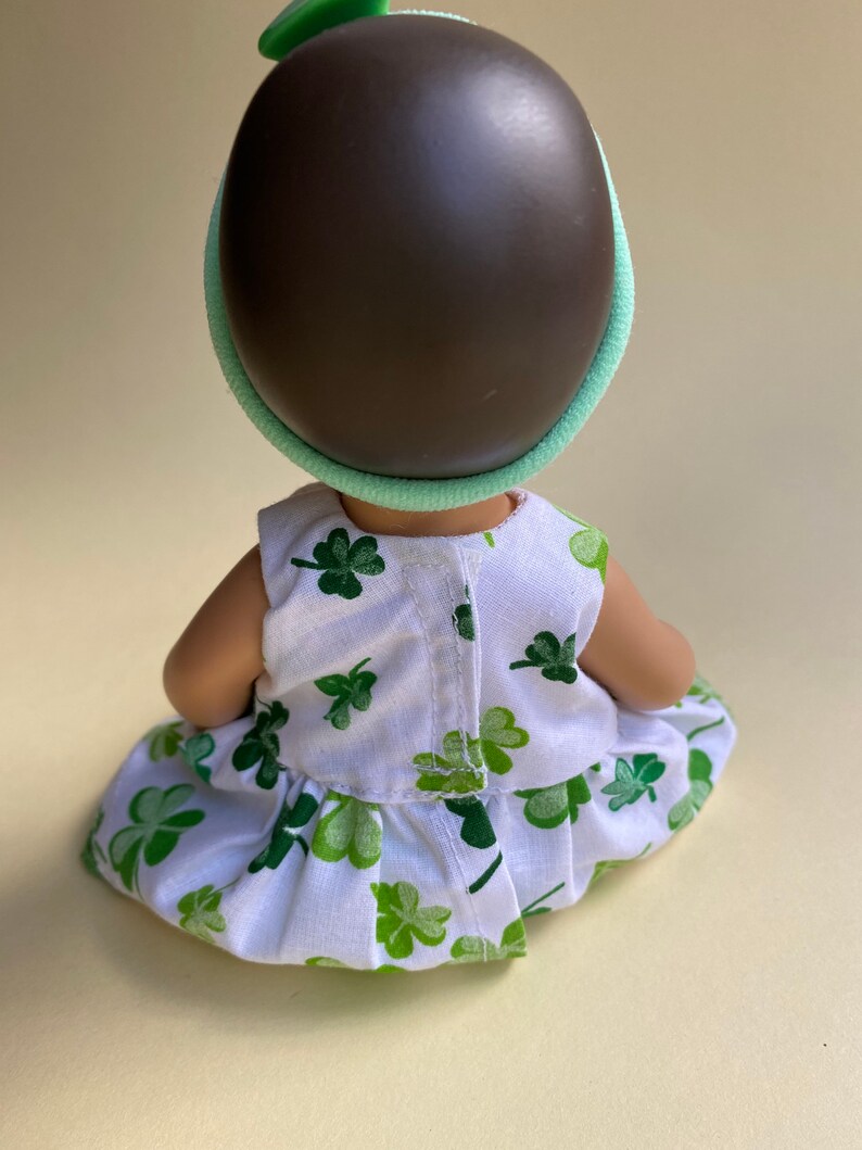 May include: A small doll wearing a white dress with green shamrock print and a green headband with a shamrock on top.