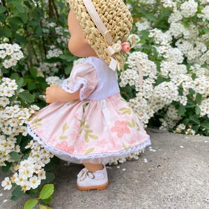 May include: A doll wearing a straw hat with a cream-colored ribbon and a pink floral dress. The dress has a white lace trim. The doll is wearing white shoes with a brown sole. The background is a lush green bush with white flowers.