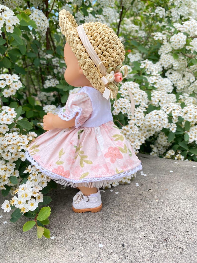 May include: A doll wearing a straw hat with a cream-colored ribbon and a pink floral dress. The dress has a white lace trim. The doll is wearing white shoes with a brown sole. The background is a lush green bush with white flowers.