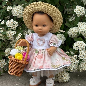May include: A doll wearing a straw hat, a pink floral dress, and white shoes. The doll is holding a small wicker basket filled with colorful flowers. The dress has a white bodice with a heart design. The doll is standing in front of a backdrop of white flowers.