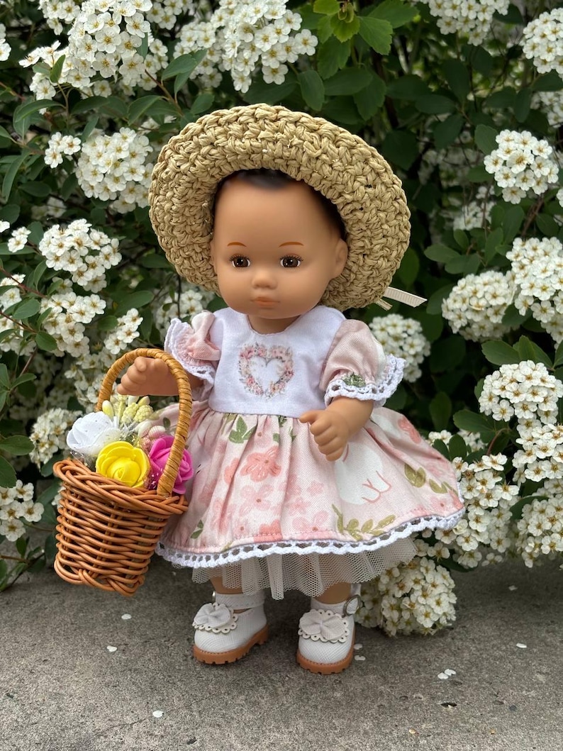 May include: A doll wearing a straw hat, a pink floral dress, and white shoes. The doll is holding a small wicker basket filled with colorful flowers. The dress has a white bodice with a heart design. The doll is standing in front of a backdrop of white flowers.