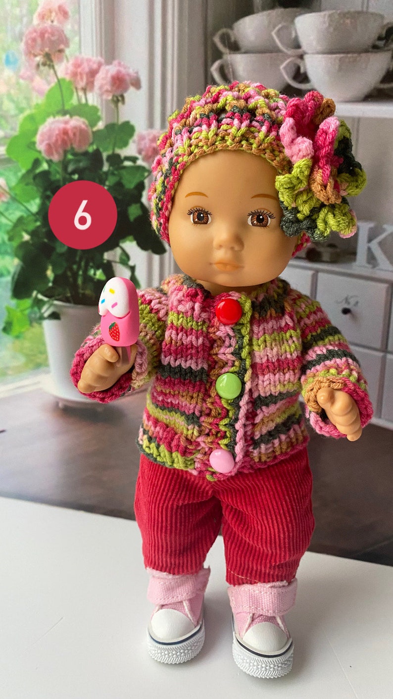 Puede incluir: Una mu&ntilde;eca con un c&aacute;rdigan de punto a rayas rosa, verde y marr&oacute;n con un gorro a juego y una flor rosa. La mu&ntilde;eca sostiene un helado de juguete rosa y blanco. La mu&ntilde;eca lleva pantalones de pana roja y zapatillas blancas con detalles rosas.