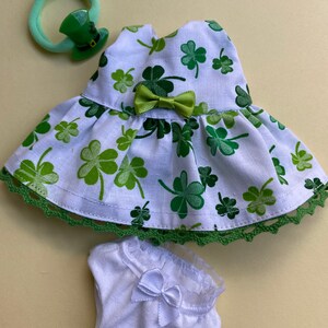 May include: A white dress with a green shamrock print and green trim. The dress has a green bow at the waist. A white pair of underwear with a white bow and lace trim. A green hair tie with a green leprechaun hat charm.