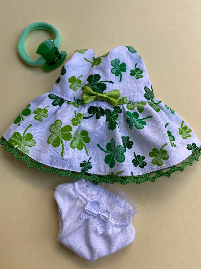 May include: A white dress with a green shamrock print and green trim. The dress has a green bow at the waist. A white pair of underwear with a white bow and lace trim. A green hair tie with a green leprechaun hat charm.