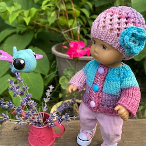 Puede incluir: Una mu&ntilde;eca con un gorro de punto rosa y morado con una flor azul, un c&aacute;rdigan de punto a rayas rosa y azul, pantalones de pana rosa y zapatillas blancas. La mu&ntilde;eca est&aacute; mirando una mariposa de juguete azul y morada.
