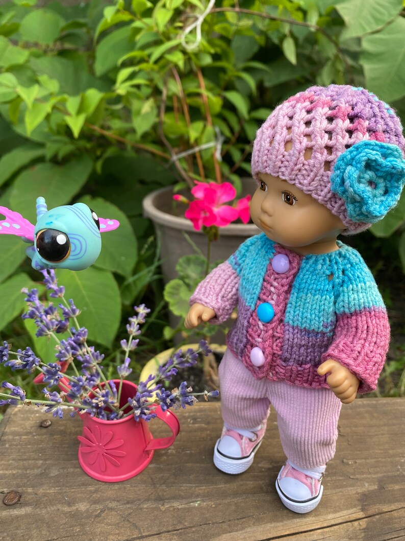 Puede incluir: Una mu&ntilde;eca con un gorro de punto rosa y morado con una flor azul, un c&aacute;rdigan de punto a rayas rosa y azul, pantalones de pana rosa y zapatillas blancas. La mu&ntilde;eca est&aacute; mirando una mariposa de juguete azul y morada.