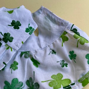 May include: White cotton fabric with a green shamrock print. The fabric has a ruffled edge and is sewn with a white thread.