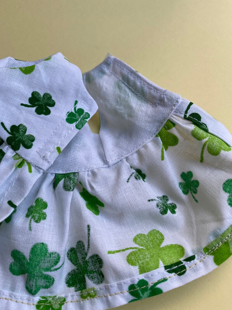 May include: White cotton fabric with a green shamrock print. The fabric has a ruffled edge and is sewn with a white thread.