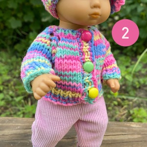 Puede incluir: Una peque&ntilde;a mu&ntilde;eca que lleva un su&eacute;ter de punto a rayas rosa y azul, pantalones rosas y zapatillas blancas. La mu&ntilde;eca lleva un gorro de crochet de colores arco&iacute;ris. La mu&ntilde;eca est&aacute; de pie sobre una superficie de madera con dos peque&ntilde;os gatos de juguete.
