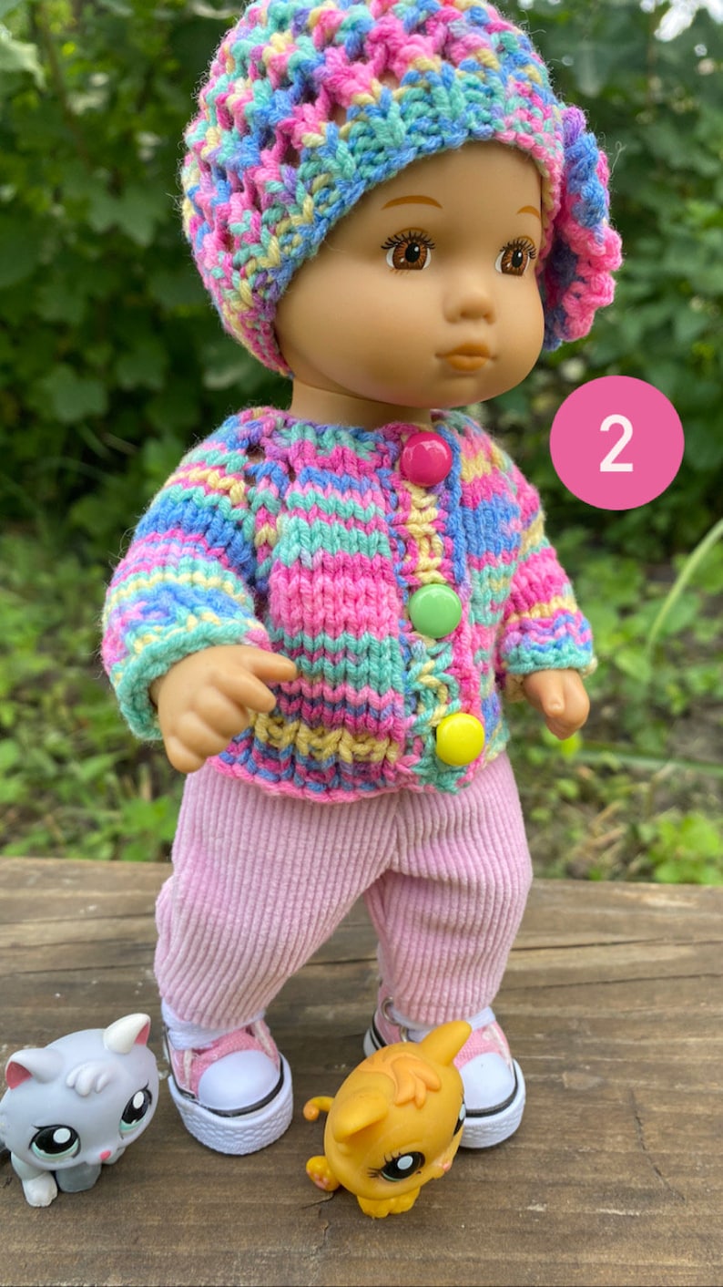 Puede incluir: Una peque&ntilde;a mu&ntilde;eca que lleva un su&eacute;ter de punto a rayas rosa y azul, pantalones rosas y zapatillas blancas. La mu&ntilde;eca lleva un gorro de crochet de colores arco&iacute;ris. La mu&ntilde;eca est&aacute; de pie sobre una superficie de madera con dos peque&ntilde;os gatos de juguete.