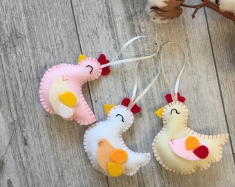 May include: Three felt chicken ornaments with white string hangers. The chickens are pink, white, and yellow with red beaks and black eyes.