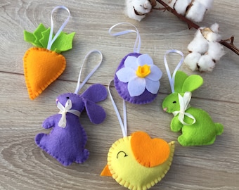 May include: Five felt Easter decorations with hanging loops. A purple bunny, a green bunny, a yellow chick, a white flower with a yellow centre, and an orange carrot.