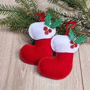 May include: Two red felt Christmas stockings with white felt cuffs and green holly accents. The stockings are decorated with red glitter berries.