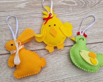 May include: Three felt ornaments, a yellow chick, a green chicken, and an orange bunny, with white ribbon bows. The ornaments are hanging from white string loops.