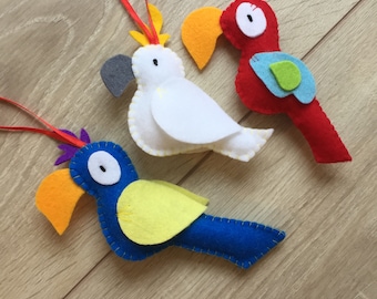 May include: Three felt bird ornaments, a white cockatoo, a red parrot, and a blue parrot. Each bird has a yellow beak and a black eye. The birds are hanging from red, white, and blue ribbons.