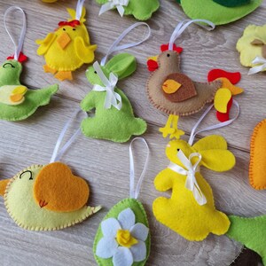 Puede incluir: Una colección de coloridos adornos de fieltro de Pascua. Los adornos incluyen pollitos, patos, conejos, una gallina y un huevo con una flor. Cada adorno tiene una cinta blanca para colgar. Los adornos son en tonos de amarillo, verde, marrón y naranja.