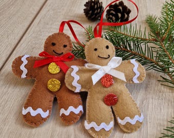 May include: Two felt gingerbread person ornaments with stitched details. One is dark brown with a red ribbon and gold glitter buttons, the other is light tan with a white ribbon and red glitter buttons. Both have white wavy trim and red hanging loops.
